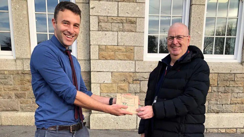 History teacher Alex Morgan (left), is wearing a blue shirt, brown tie and jeans. On the right is ex-student Sean Morris who is wearing a black coat 
Behind them is the brick of the school. Mr Morgan is holding out an old exercise book to Mr Morris