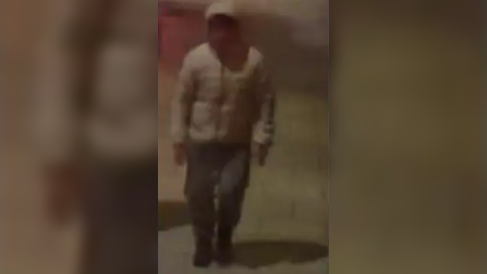 A blurry CCTV image showing a man walking down the street wearing a light grey puffed jacket with his hood up, dark trousers and black shoes