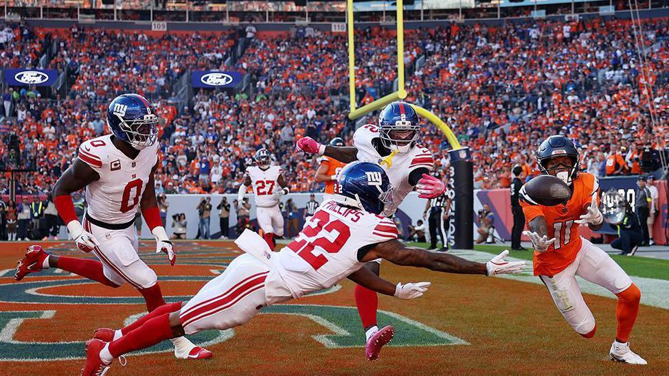 American football player in orange catches the ball near the end zone as three defenders in white and red dive and reach to block during a crowded stadium game.