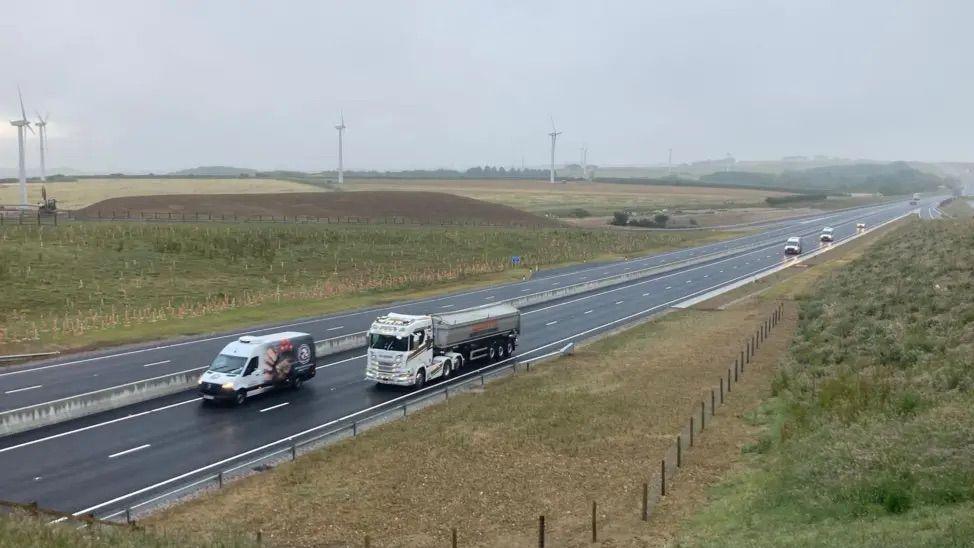 A dual carriageway with cars and lorries driving on it. There are wind turbines to the left of the dual carriageway.