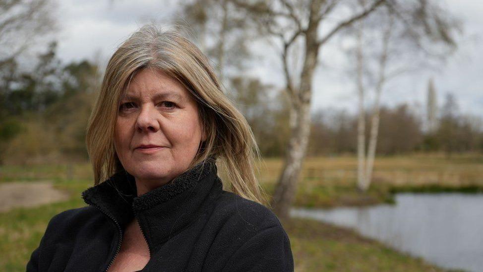 Alison Downes smiles at the camera. She is standing in a field with a pond behind her. She has long blonde hair and is wearing a black jumper