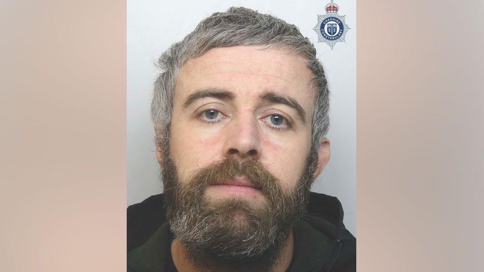 A police mug shot of Lee Bailey, he has short grey hair and a scruffy brown and grey beard. He wears a dark hoodie.