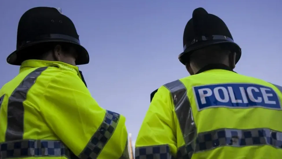 Stock image of the backs of police officers against a blue sky. They are wearing black hats and hi-vis jackets which say the word "Police". 