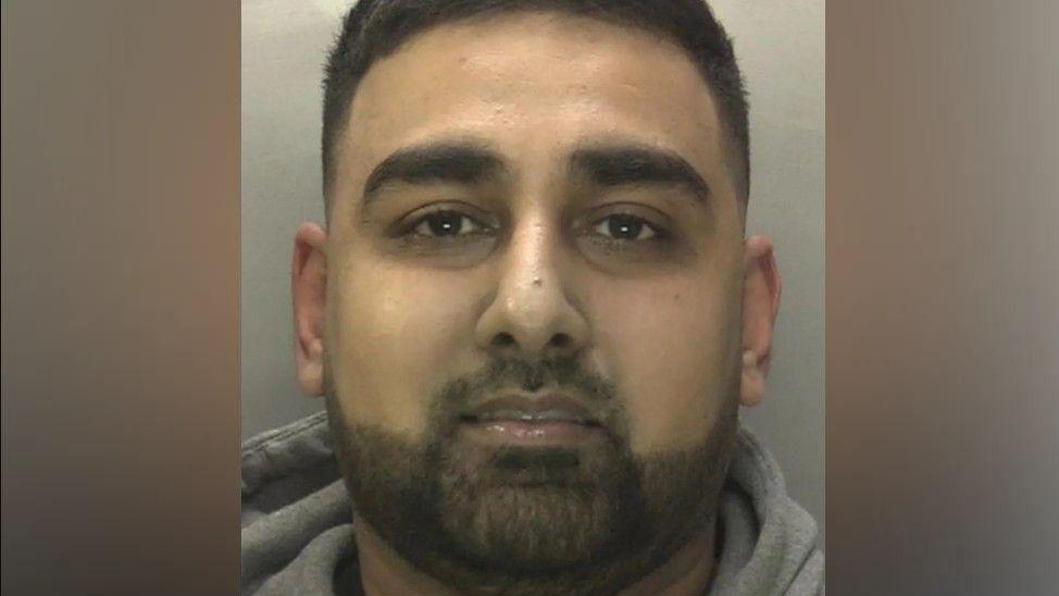 A mugshot of a man staring at the camera. He is wearing a grey hoodie.