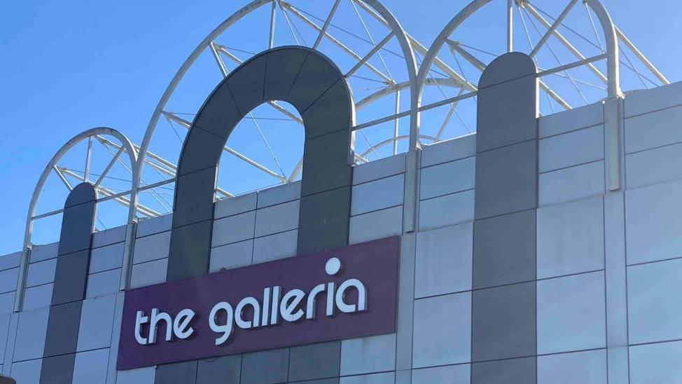 The front of a white-and-grey building with a purple rectangular which has the words "the galleria" written in white, lower-case, letters