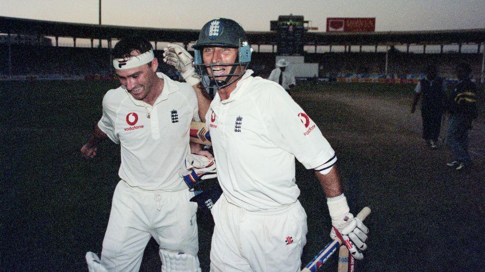 Two men wearing cricket whites smiling and walking towards the camera as they exit a cricket pitch in a stadium.
