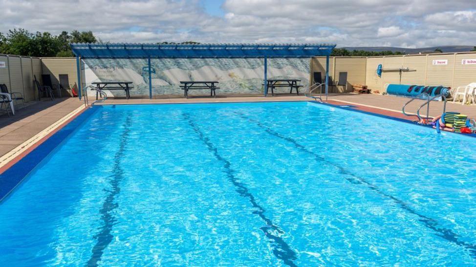 Lengthways view of a swimming pool full of water, but with no swimmers. There are benches on a partially-covered paved area at one end, and to the left a rolled up blue tarpaulin cover and some swimming aids.