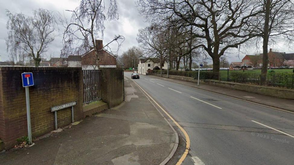 A Google street view image of a road going past a school with a the boundary fence and wall visible on the right. On the left is a wall with a dead end sign and a street sign which says "union street".