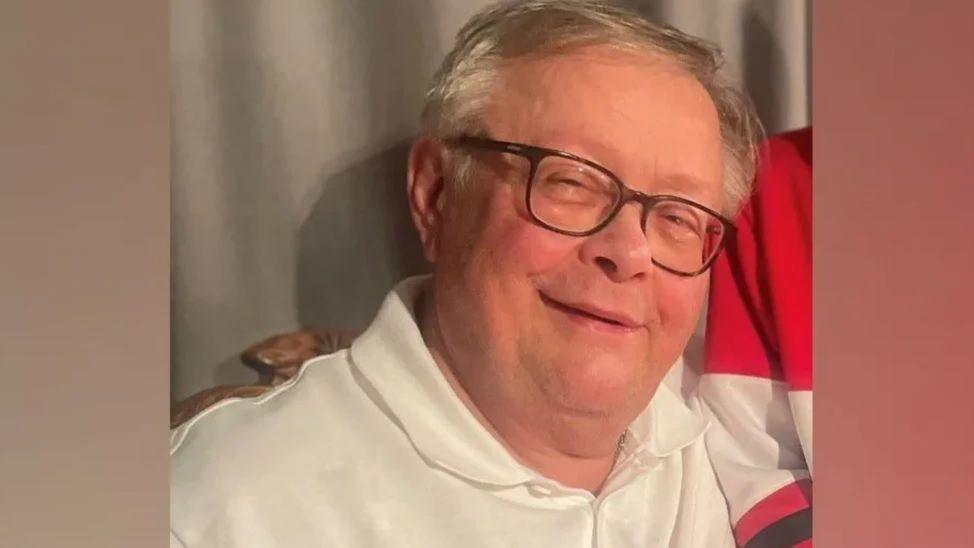 A family-issued photograph of Stuart Everett. He has white hair, has glasses on, and is smiling wearing a white polo shirt.