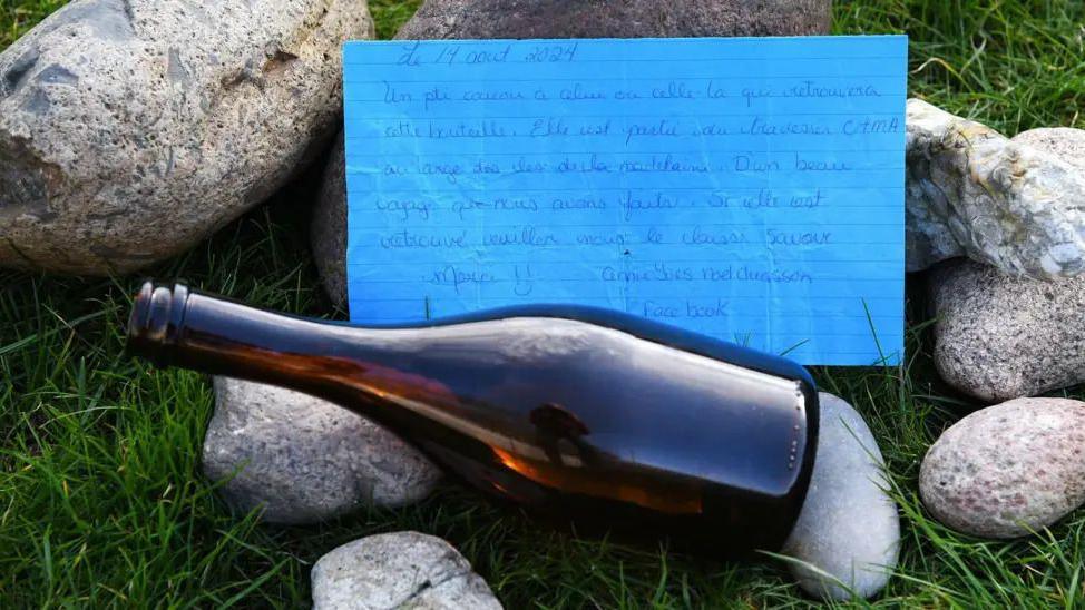 a brown bottle laid on some grass and stones with a letter written on blue paper above it.
