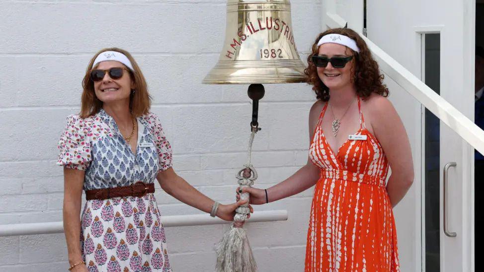 A mother and daughter both ringing a large bell attached to a white brick wall.