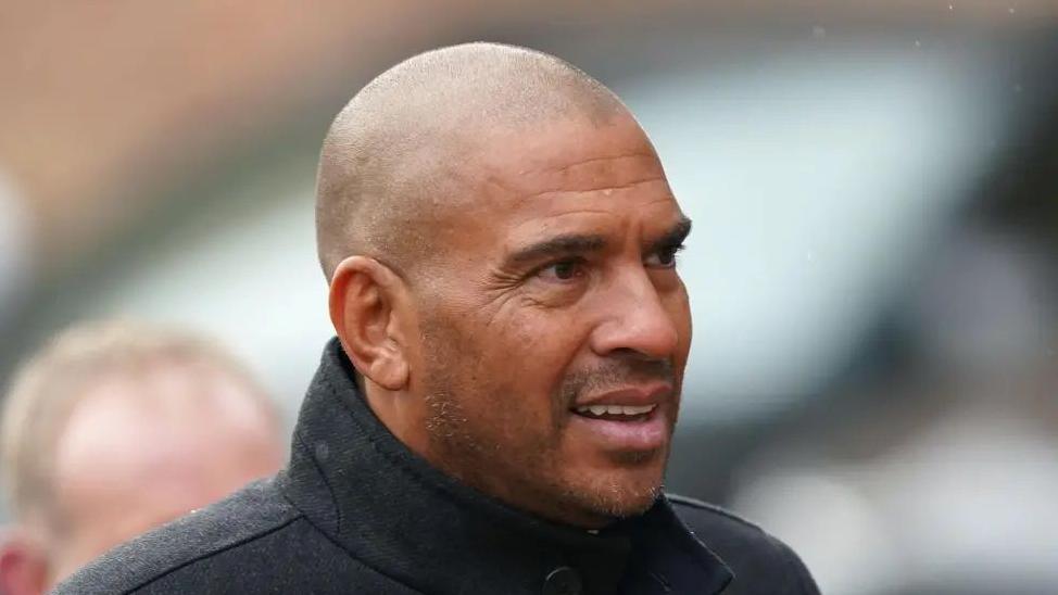 Stan Collymore with very short dark hair looking to the right of the camera while wearing a black coat. He is in a crowd of people, possibly at a football match.
