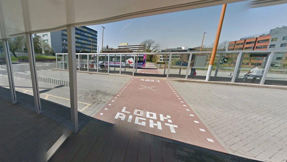 A screenshot from Google street view of a bus station. It's a large forecourt with bus shelters on the pavements with glass walls. There are tall buildings in the background.