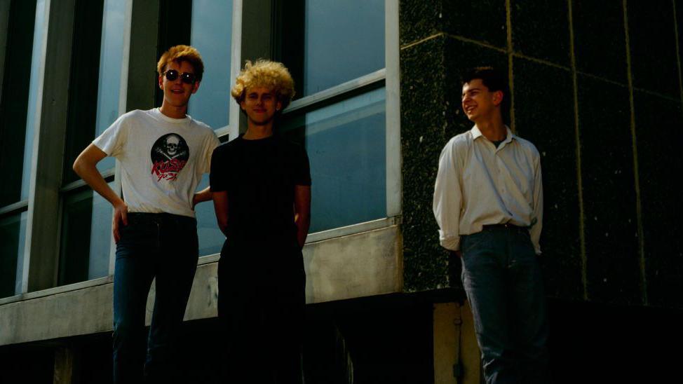 Andy Fletcher stands with his hands on his hips and smiling at the camera, next to Martin Gore, who is wearing all black and has curly blonde hair and Dave Gahan, who is wearing a shirt and jeans and looking at the other two and smiling at them. They are standing in front of a public building, made of steel, glass and stone concrete panels.
