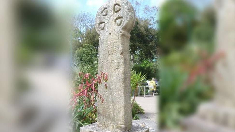 The picture shows an old granite cross mounted on a stone base. The cross has a circular head with four triangular recesses arranged around the centre. The surface appears weathered. It is set outdoors in a landscaped area with plants nearby, and there are tables and chairs in the background.