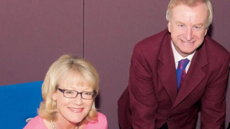 Wendy Austin sitting at a desk smiling up at the camera. She is wearing a pink top. Seamus McKee is standing leaning over with his hands over the desk. He is wearing a suit and tie.