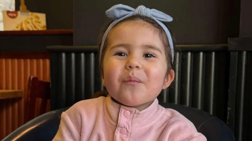 Louisa "Lulu" Palmisano, three, wearing a pink fleece and a blue bow in her brown hair, sits on a black leather chair in a restaurant and smiles at the camera.