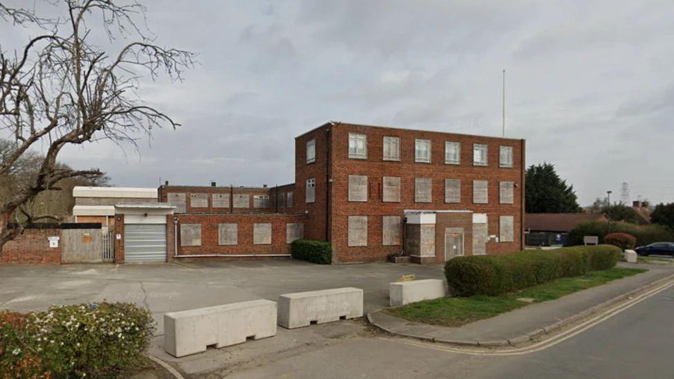 An exterior shot of a detached red brick office building  with a hedge pavement and road in foreground.