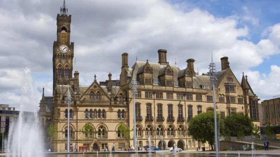 A grand Victorian gothic building with a clock tower on the left end of the building. An open paved space in front of the building with a fountain on the left and three poles with lights at the top are to the right of the fountain.