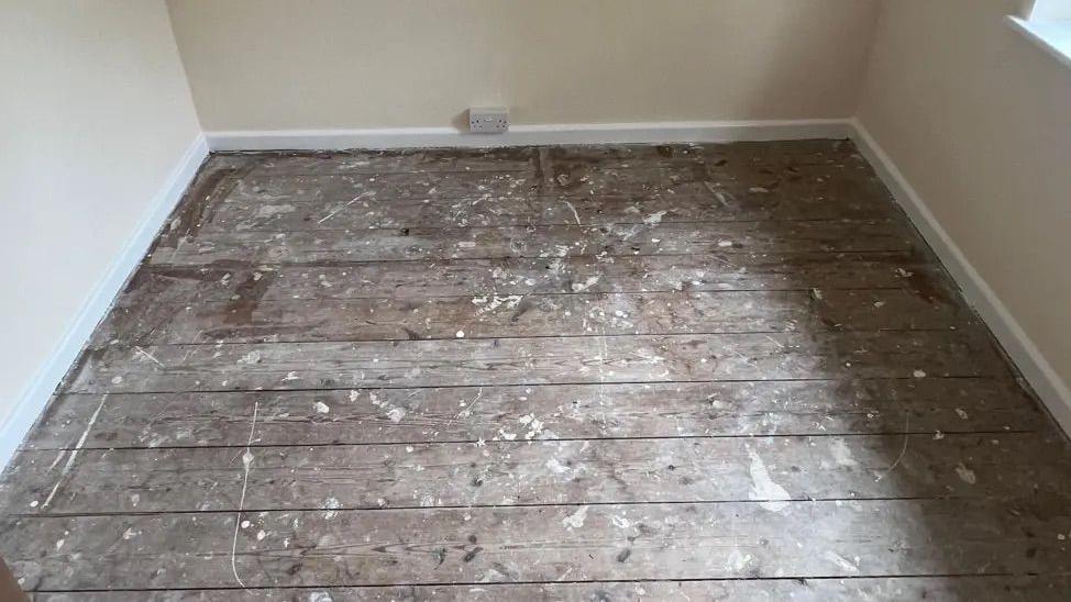 Empty room with bare floorboards. The wall is cream. There are white rims around the floorboard. There is an outlet on the wall. 