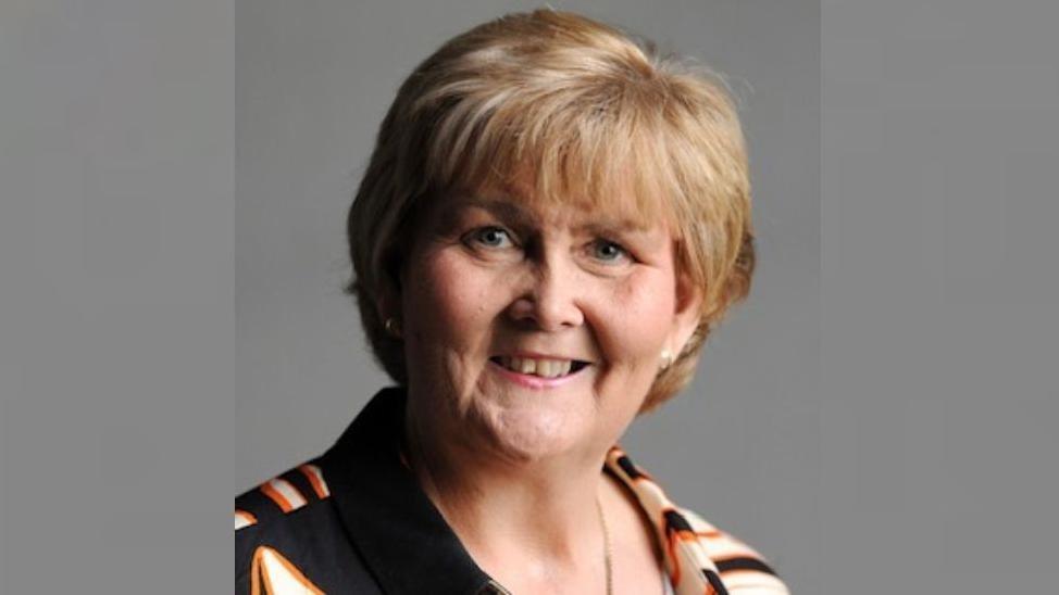 Passport-photo style image of Tracey Dixon who is smiling. She has a bob of blonde hair and is wearing a striped shirt with a chain. The stripes on the shirt are black, orange and white.