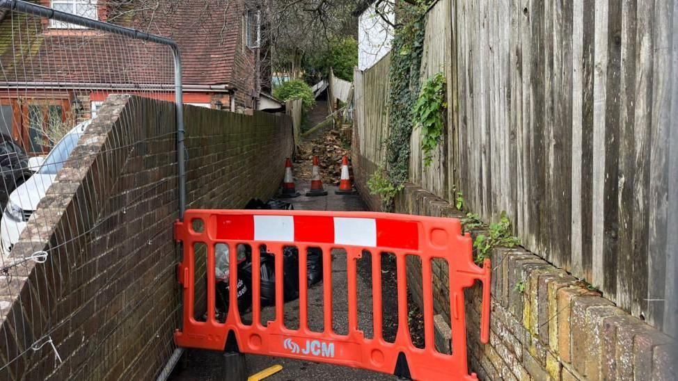 The narrow path with walls on either side which is blocked by a barrier and cones and further down a pile of what looks like rubble blocking the way.