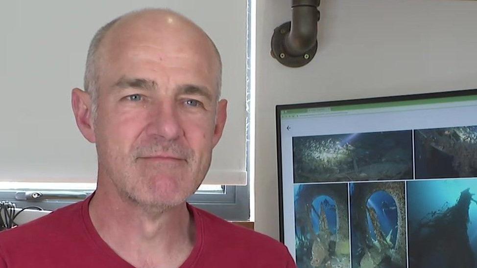 Dom Robinson - wearing a red T-shirt and has grey hair and a bald head. A computer screen with diving footage is on the screen behind him