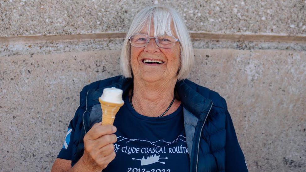 A woman with grey hair and glasses in a blue T-Shirt and top smiles while eating an ice cream cone