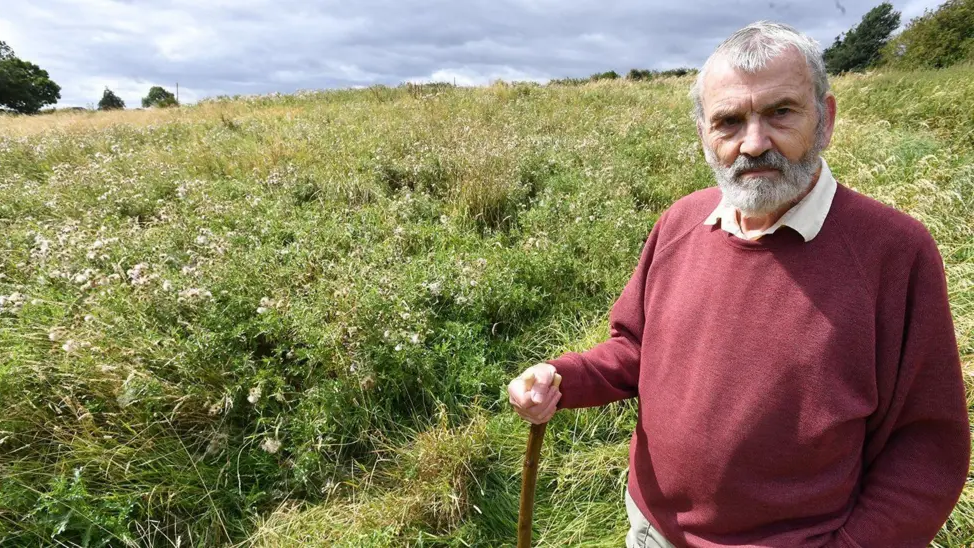 Robert Smith, who has grey-haired and a beard and is wearing a maroon jumper and a serious expression, is holding a walking stick or staff, with a field of overgrown thistles and grass in the background.