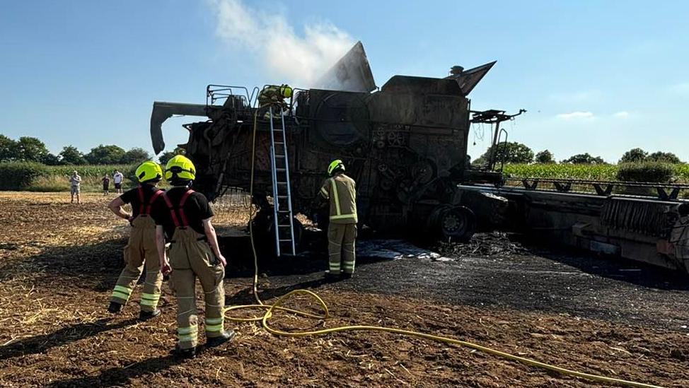 Three firefighters standing in a field next to a burnt-out combine harvester. There is a ladder leaning against the combine harvester with another firefighter standing on its roof. The ground surrounding the farm machinery is black.