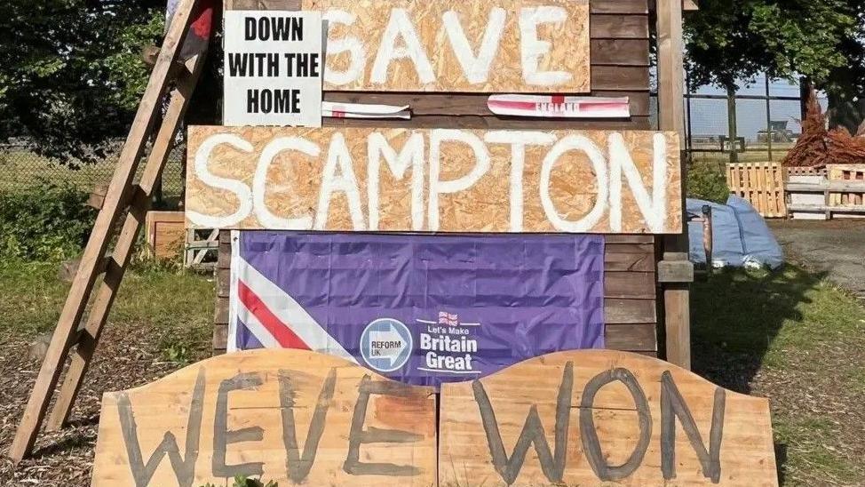 A "Save Scampton" sign on a wooden backing outside the former RAF site. Another sign at ground level reads: We've Won". There is a set of ladders leant up against the display.