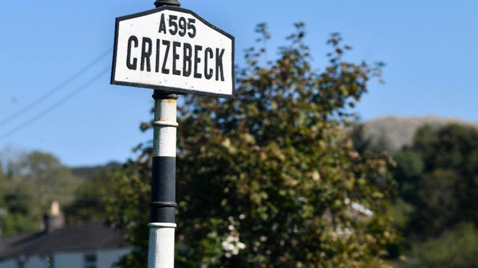 An old-fashioned sign for Grizebeck on a black and white post with some trees and a white house behind