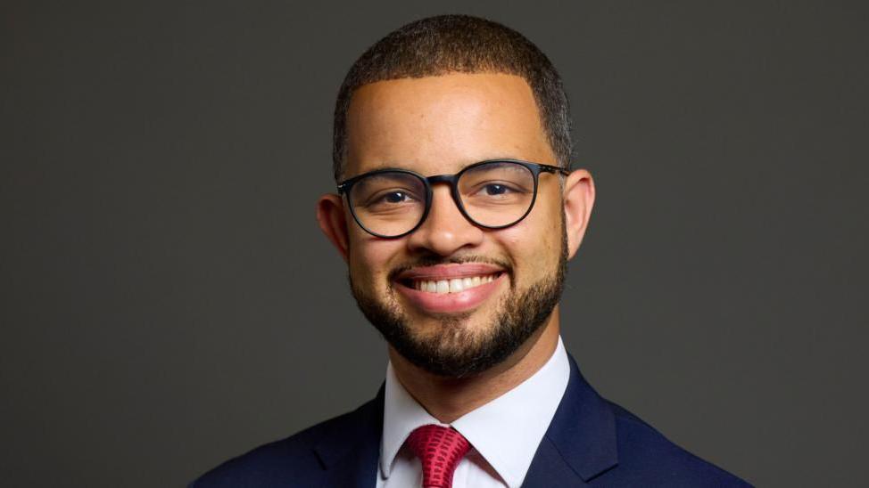 Adam Jogee wearing glasses, a suit and red tie, smiles at the camera in front of a dark grey background.
