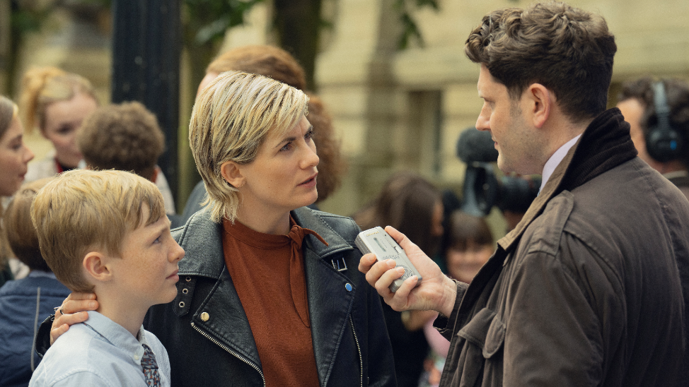 A still picture from a scene in the Toxic Town drama. Jodie Whittaker, in a black leather jacket and brown top, has her arm around a young boy. They are being interviewed by a male reporter in a brown coat who is holding a tape recorder towards Jodie Whittaker. They interview is taking place outside a court building.