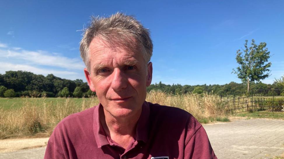 Martin Baker looks directly at the camera, dressed in a red polo neck shirt and with meadows behind him. 