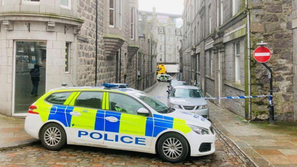 Police car with yellow and blue checks on a cobbled street, with blue and white police tape across a street.