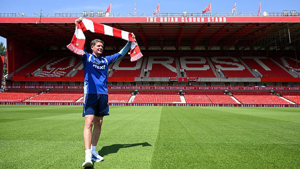 Oliver Glasner holding a Nottingham Forest scarf above his head on the pitch at an empty City Ground. The Brian Clough Stand is behind him.