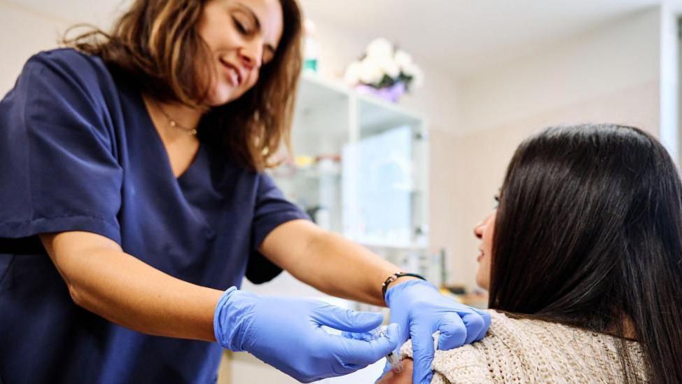 A woman dressed in a blue hospital uniform and wearing blue plastic gloves gives an injection to a woman by sticking a needle in her left upper arm. 