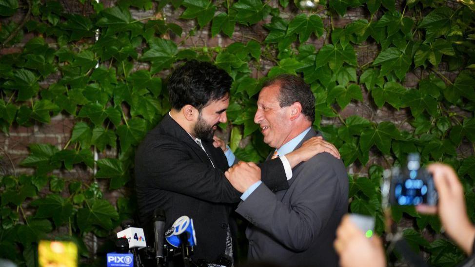 Mamdani is dressed in a dark suit as he embraces Lander who is wearing a grey suit, light blue shirt and coloured tie. They are standing in front of a brick wall covered in green leaves and they are looking intently at each other smiling