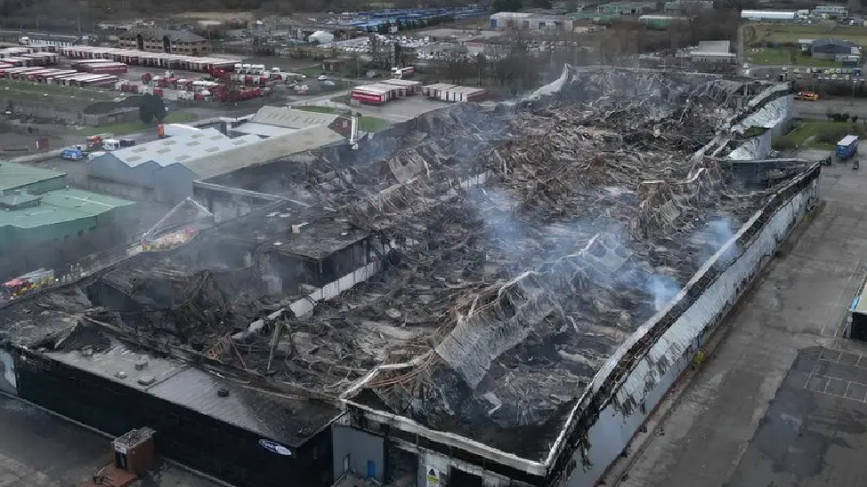 An aerial image showing the extensive damage of the fire to the large warehouse. There is still smoke blowing from the collapsed roof. Fire trucks can be seen behind the warehouse.