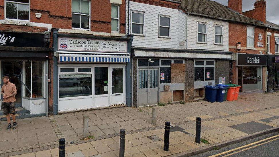 The street scene before demolition work was carried out, showing a shop front with boards across some windows and whitewash on others.