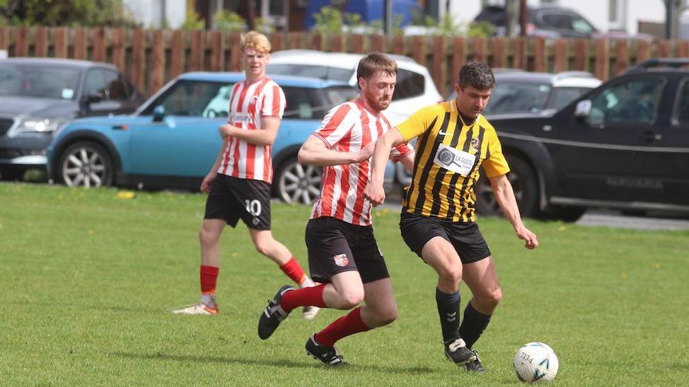 A man with a yellow and black stripy t-shirt is kicking a ball, and Kian is about to tackle him with a red and white stripy top, another player with a red and white stripy top is in the background watching the tackle.
