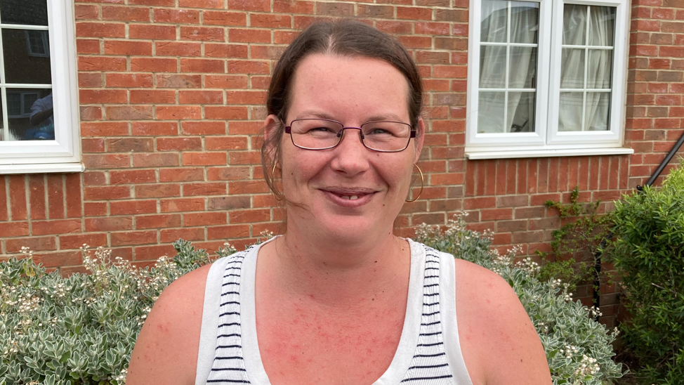 Ms Weise is standing outside a red bricked house and smiling. She is wearing rectangular framed glasses and a black and white striped vest top. She has brown hair that has been tied back away from her face and hoop earrings. 