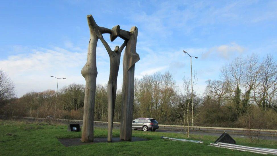 Wooden arch depicting a human figure with their arms held up, surrounded by a wooden frame. It sits on grass with blue sky above and a road with a car driving in the background. 