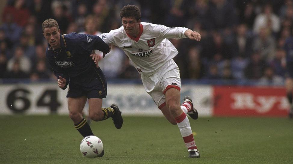 Wimbledon's Chris Perry (left) holds off Niall Quinn as they chase the ball. Perry is wearing an all-navy strip with yellow edging, while Quinn is in white with red detailing.