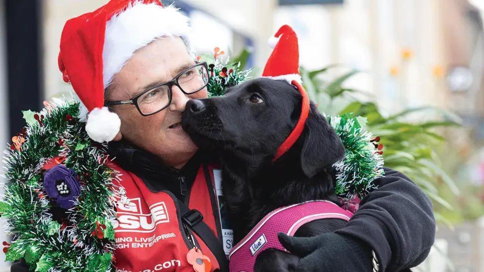 Nick Cuthbert, in Big Issue clothing, hugging Bracken, a black labrador. Nicky has glasses and is wearing a red Christmas hat. Bracken is wearing a headband with a Christmas hat. Bracken is also in a pink harness. Both have tinsel around them.