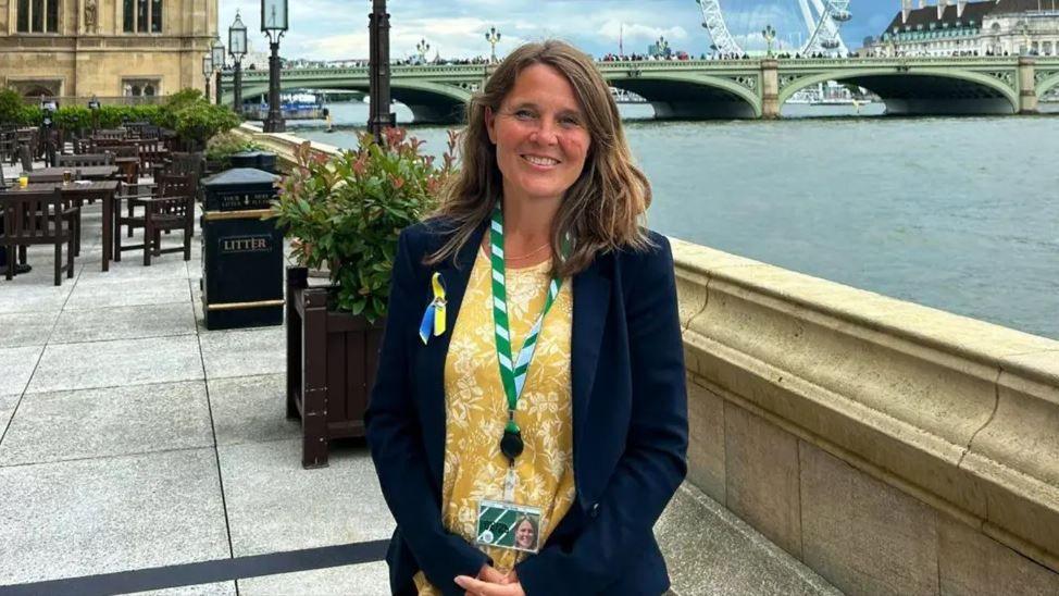 Vikki Slade stands outside with the London Eye in the background and a bridge over the River Thames. She is smiling at the camera, has mid length light brown hair and wears a navy blue blazer with a yellow and white floral print top underneath.