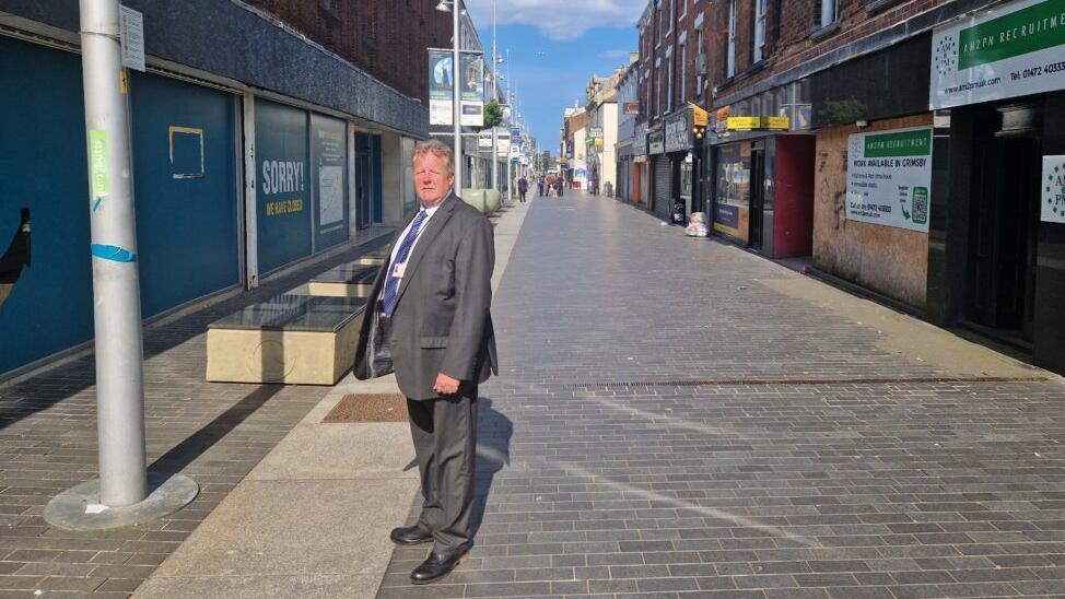 Councillor Ron Shepherd, wearing a grey suit, stands in a pedestrian street in Grimsby town centre, next to a large, grey post designed to house a tannoy system. 