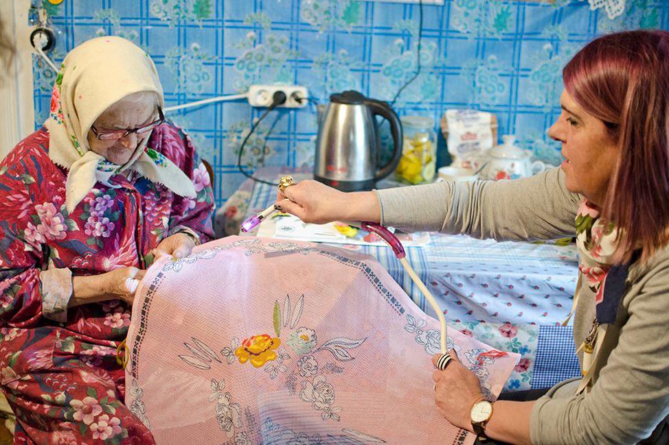 Claire Baker stitching with grandmother Baba Hana at a small kitchen table. The wall has a bright blue square pattern and Baba Hana, wearing cream headscarf and dark pink floral dress, is looking down at her work. There is a kettle and tea pot on the table.