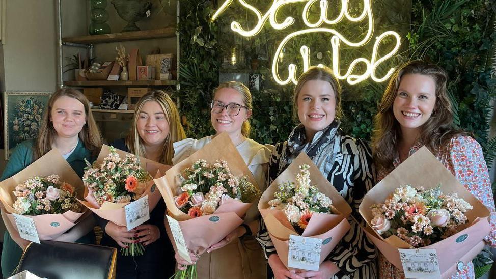 Five women each holding pink and cream bouquets of flowers smiling at the camera. They are in a florists studio. 